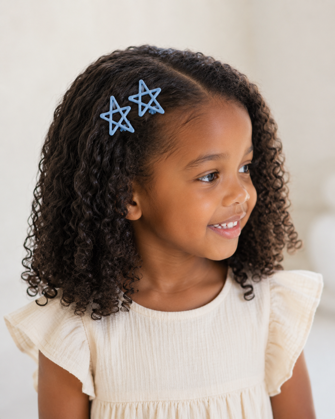 A young girl with curly dark brown hair wearing two light blue star-shaped hair clips, dressed in a cream-colored ruffled top against a soft, light beige background.