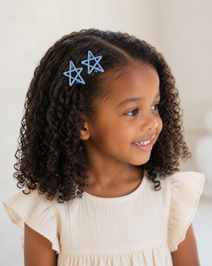A young girl with curly dark brown hair wearing two light blue star-shaped hair clips, dressed in a cream-colored ruffled top against a soft, light beige background.