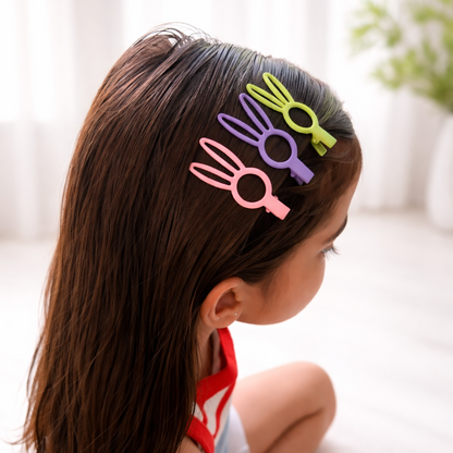 Three bunny-shaped hair clips in pink, purple, and green shown on a child's brown hair in a softly lit indoor setting.