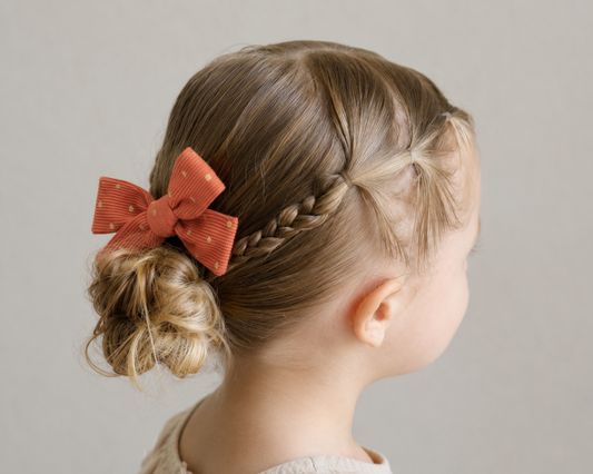 A young girl with light brown hair styled in a half-up braid with a twisted bun and a large terracotta orange bow, shown from the back against a neutral gray background.