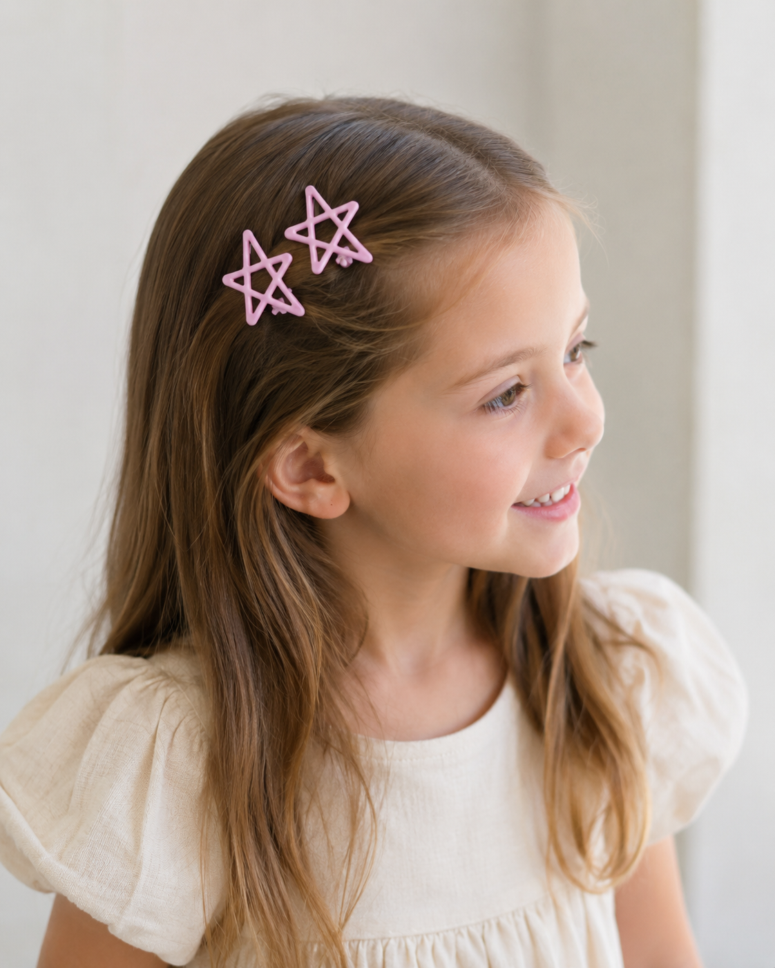 A young girl with long brown hair wearing two pink star-shaped hair clips, shown in a close-up portrait against a soft light background.