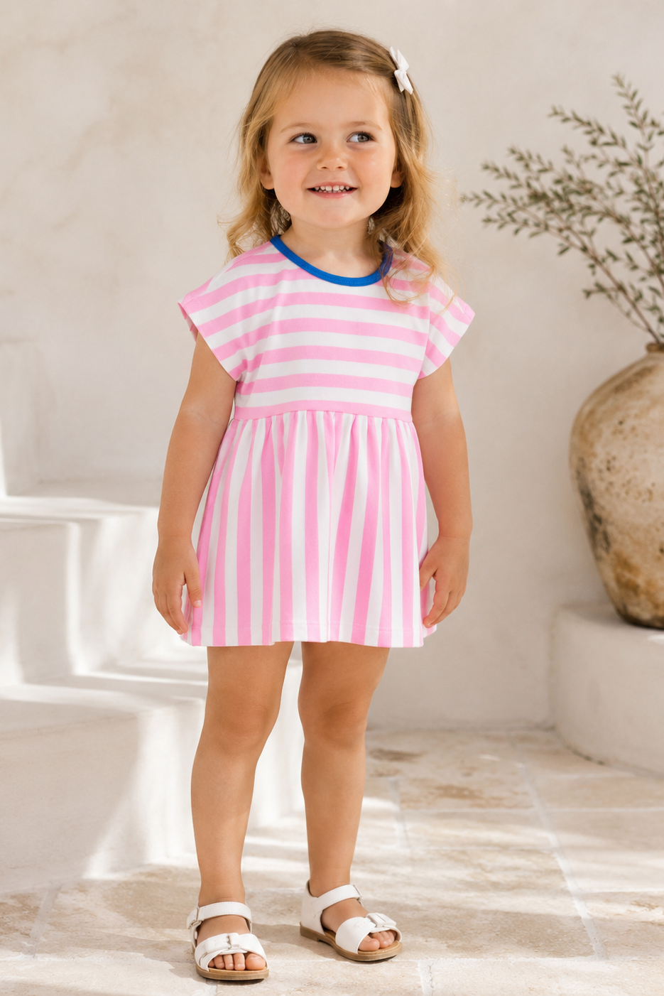 Young girl wearing a pink and white striped dress standing in a room with a vase and plant in the background.