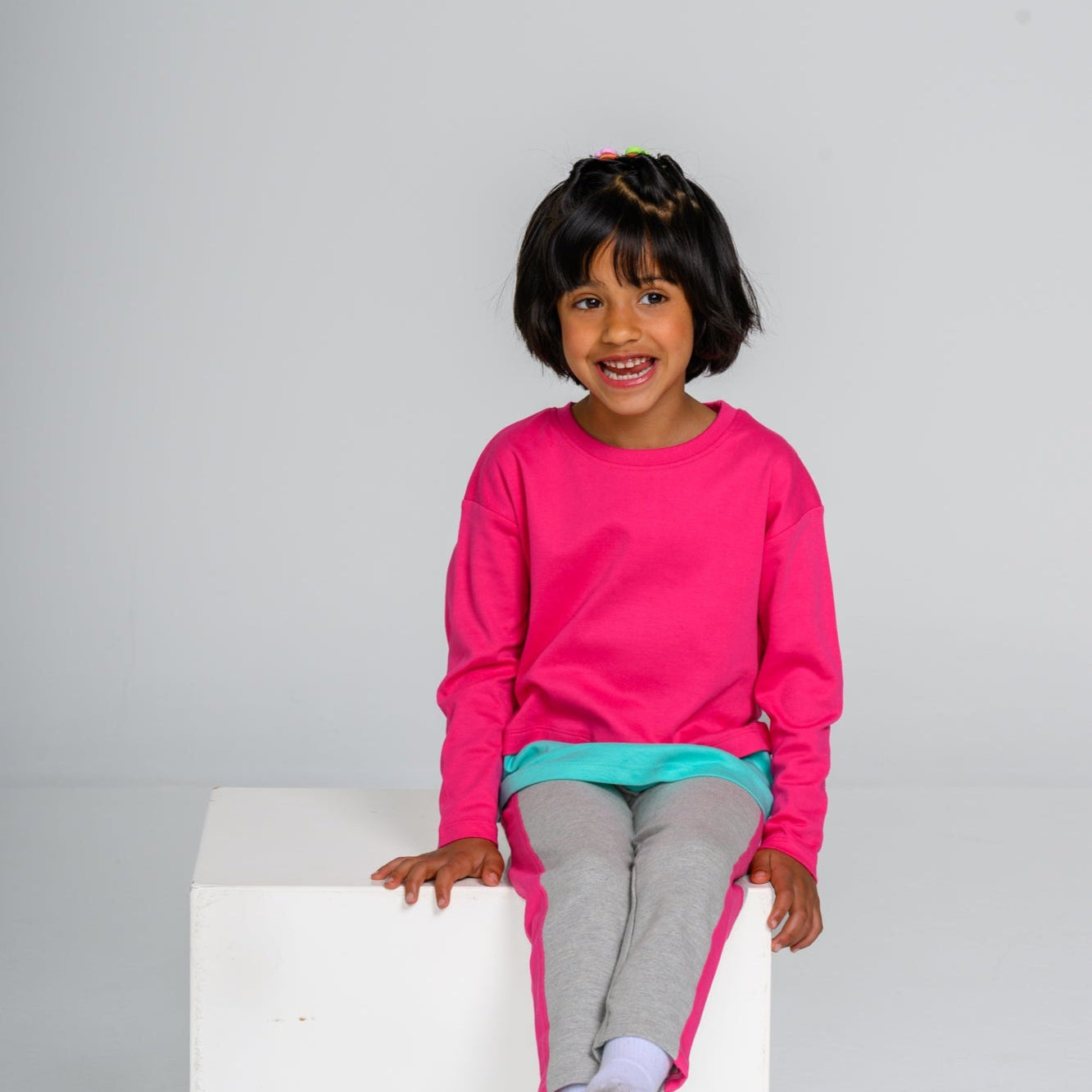 Child wearing a bright pink sweatshirt and gray pants sitting on a white box against a plain background