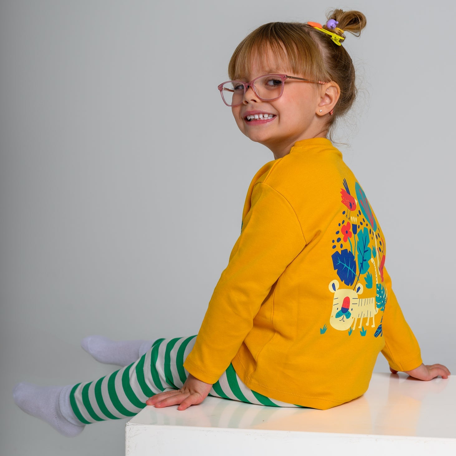 Young girl wearing a yellow sweater with a colorful design, sitting on a white surface against a gray background.