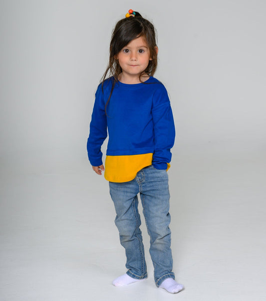 Child wearing a blue color block tshirt with a yellow layer and blue jeans on a plain background