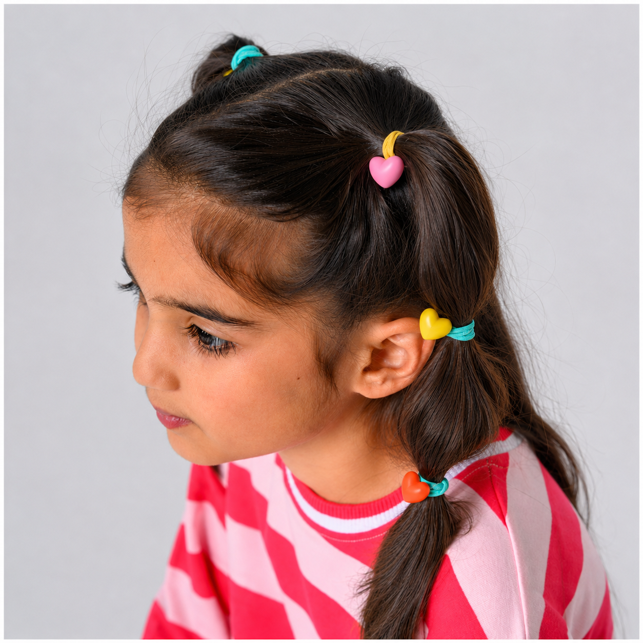 A young girl with dark brown hair styled in two buns and a side braid, wearing colorful heart-shaped hair ties in pink, yellow, teal, and orange. She is wearing a pink and white striped shirt against a solid light gray background.
