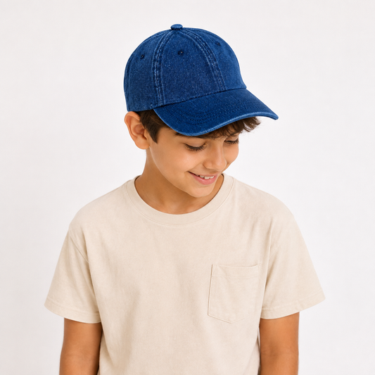 A young boy wearing a blue denim baseball cap and a light beige crewneck t-shirt, looking down and smiling against a plain white background.