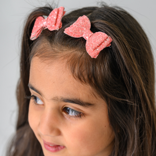 A close-up of a young girl with dark brown hair wearing two pink glittery bow hair clips, shown against a neutral light gray background.